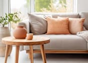 Thumbnail 3 of 11 - A living room with a grey couch, a round wooden table with a vase and candle on it, and a potted plant.