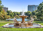 Thumbnail 41 of 44 - A fountain in the middle of a park with trees and buildings in the background.