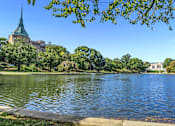 Thumbnail 40 of 44 - A serene lake surrounded by lush greenery and a building with a green spire in the background.