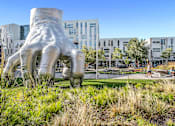Thumbnail 33 of 44 - A large sculpture of a hand is in the foreground of a grassy area with buildings in the background.