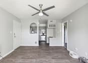 Thumbnail 6 of 32 - A room with a ceiling fan and a kitchen area in the background.