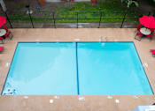 Thumbnail 14 of 28 - A rectangular pool with a blue tint is surrounded by a black fence and has red umbrellas on the side.