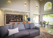 Thumbnail 29 of 32 - A grey couch with a white pillow is in the middle of a room with a bar area in the background.