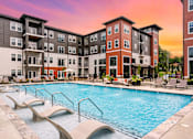 Thumbnail 1 of 43 - A large swimming pool is surrounded by lounge chairs in front of apartment buildings.