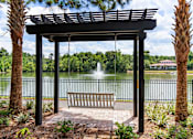 Thumbnail 42 of 50 - A wooden bench is under a pergola next to a lake.