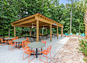 Thumbnail 24 of 50 - A wooden pavilion with orange chairs and a green table is surrounded by trees.