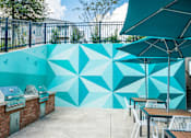 Thumbnail 20 of 31 - A wall with a geometric mural is behind a row of tables and grilling area.