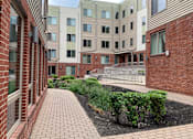 Thumbnail 4 of 11 - A brick building with a walkway and bushes in front.