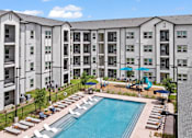 Thumbnail 3 of 19 - A large swimming pool surrounded by lounge chairs and umbrellas in front of a multi-story apartment building.