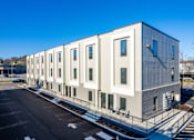 Thumbnail 15 of 23 - A modern building with a grey and white exterior and a parking lot in front.