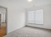 Thumbnail 24 of 31 - A room with a carpeted floor, a window with blinds, and a door leading to another room.