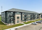 Thumbnail 3 of 26 - A large grey building with a parking lot in front.