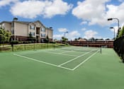 Thumbnail 50 of 64 - A tennis court is surrounded by a black fence and a building is in the background.