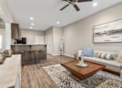 Thumbnail 6 of 11 - 2-Bedroom Senior Townhomes in Belton, MO - Traditions Villas 55+ - Living Room with Vinyl Plank Flooring, High Ceiling, Ceiling Fan, Couch, Coffee Table, Painting, Large Rug, and a View of the Kitchen