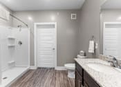 Thumbnail 11 of 22 - Shawnee KS Pet-Friendly Luxury Apartments - Bonavia Luxury Apartments - Bathroom with Vinyl Plank Flooring, Vanity Mirror, Sink, Dark Brown Cabinetry, and Open Shower.