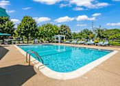 Thumbnail 14 of 30 - A large outdoor swimming pool surrounded by trees and a clear blue sky.