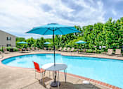 Thumbnail 8 of 23 - A blue umbrella is next to a table with chairs by a pool.