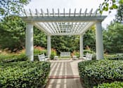 Thumbnail 21 of 28 - A white pergola with benches is surrounded by green bushes.