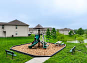 Thumbnail 13 of 47 - Townhomes in Peculiar, MO - Bradley’s Crossing - An Outdoor Playground Featuring a Play Structure on a Wood Fiber Surface Surrounded by Lush Grass, Trees, Benches, and Townhomes.