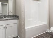 Thumbnail 11 of 23 - Three-Bedroom Townhomes in Belton, MO - Traditions Townhomes - Bathroom with Vinyl Plank Flooring, Built-in Bathtub, Single Vanity, Sink, Mirror, and White Cabinetry.