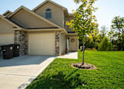 Thumbnail 4 of 23 - 2-Bedroom Townhomes in Belton, MO - Traditions Townhomes - A Garage Attached to the House, with Driveway, Black Garbage Bins, and Lush Landscaping.