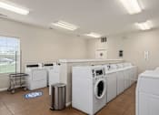 Thumbnail 12 of 28 - Dog-Friendly Apartments in Lawrence, KS - Remington Square - Laundry Room with a Washer, Numerous Dryers, Trash Bin, Laundry Cart, Light Fixtures, and a Large Window with Blinds.