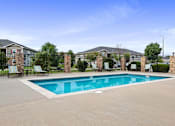 Thumbnail 22 of 28 - Pet-Friendly Apartments in Lawrence, KS - Remington Square - Swimming Pool with a Spacious Sundeck, Surrounded by Trees, Black Metal Fence, and Lounge Chairs.