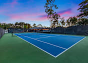Thumbnail 21 of 22 - A tennis court with a blue surface and white lines is surrounded by a fence and trees.