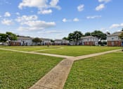 Thumbnail 15 of 19 - A park with a pathway in the middle of a grassy area with buildings in the background.