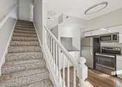 Thumbnail 4 of 27 - A staircase with a carpeted runner and white balusters leads to a kitchen with stainless steel appliances.
