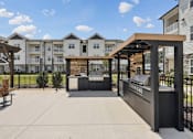 Thumbnail 4 of 26 - A sunny day at a residential complex with a covered walkway and apartment buildings with outdoor kitchen and grills.
