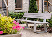 Thumbnail 145 of 149 - A grey bench is in front of a building with a tree and flowers.
