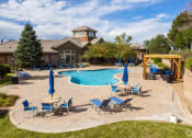 Thumbnail 10 of 32 - South Aurora, CO, Apartments for Rent - Sonoma Resort at Saddle Rock - Pool with Lounge Seats, Umbrellas, Tables, Chairs, Pergola, and Surrounding Trees.