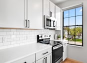 Thumbnail 39 of 53 - A kitchen with white appliances and cabinets with a window overlooking a green area.