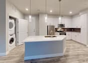 Thumbnail 21 of 43 - A modern kitchen here at Experience at Epicenter featuring white shaker-style cabinets, light countertops, and a large center island with an integrated sink and pendant lighting overhead. Stainless-steel appliances line the back wall alongside a dark tile backsplash, while an in-unit laundry closet and wood-style flooring add everyday convenience and a clean, open feel.