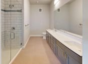 Thumbnail 27 of 43 - A modern bathroom here at Experience at Epicenter featuring light tile flooring, a long gray vanity with dual sinks, and a wide wall mirror with modern lighting. A glass-enclosed walk-in shower with white subway tile sits beside a clean-lined toilet, creating a bright, streamlined layout with ample counter space and storage.