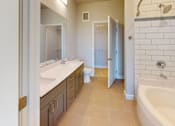 Thumbnail 28 of 43 - A spacious bathroom here at Experience at Epicenter featuring light tile flooring, a long gray vanity with dual sinks, and a wide wall mirror with modern lighting. A bathtub with white subway tile surround sits beside a clean-lined toilet, while a walk-in closet with shelving and hanging space adds convenient storage and functionality.