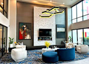 Thumbnail 35 of 43 - A modern lobby lounge here at Experience at Epicenter featuring a dramatic floor-to-ceiling fireplace wall with textured paneling, contemporary artwork, and a mounted TV. Plush curved seating, round accent tables, and a sculptural chandelier sit atop patterned carpeting, while tall windows bring in natural light and frame an inviting community gathering space.