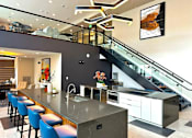Thumbnail 36 of 43 - A contemporary community kitchen and lounge here at Experience at Epicenter showcasing a large stone island with bar seating, sleek cabinetry, and built-in appliances beneath modern lighting. A glass-railed staircase leads to an upper-level seating area, while abstract artwork, polished surfaces, and open sightlines create a stylish space for gatherings and events.