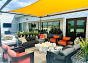 Thumbnail 38 of 43 - A stylish rooftop lounge here at Experience at Epicenter featuring a modern outdoor seating arrangement with deep gray sofas, orange accent chairs, patterned throw pillows, and a sculptural stone-style coffee table. A bright shade canopy stretches overhead, while planters, string lights, and surrounding city views create a relaxed, social atmosphere.