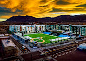 Thumbnail 41 of 43 - A dramatic aerial view here at Experience at Epicenter showcasing a vibrant urban neighborhood centered around a professional-style sports field, surrounded by modern apartment buildings and illuminated streets. The scene is framed by mountain backdrops and a glowing sunset sky, highlighting the community’s energetic, city-connected setting.