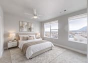 Thumbnail 7 of 43 - A spacious bedroom here at Experience at Epicenter featuring plush carpet, soft neutral walls, and an upholstered bed with layered white and beige bedding. Matching nightstands with table lamps sit beneath modern wall art, while a ceiling fan and large windows with simple shades frame scenic neighborhood and mountain views, filling the room with natural light.