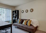 Thumbnail 12 of 32 - A bright office space here at Windsong featuring a cozy gray sofa with colorful accent pillows, sleek desk, and modern shelving. Large window with vertical blinds brings in natural light, creating a comfortable and inviting workspace.