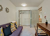 Thumbnail 13 of 32 - A versatile office space here at Windsong featuring a cozy sofa with colorful accent pillows, spacious desk, and open closet. Neutral tones and soft lighting create a comfortable, welcoming area for work or relaxation.