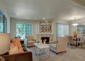 Thumbnail 4 of 32 - A bright living and dining area here at Windsong featuring a brown sofa with orange accent pillows, beige armchairs, and a mirrored fireplace. Welcoming space with soft carpet, elegant glass table, and large windows bringing natural light.