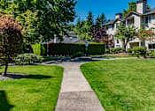Thumbnail 23 of 32 - A landscaped outdoor courtyard here at Windsong featuring a lush green lawn, mature trees, and a peaceful walking path. This bright and welcoming space offers scenic greenery and a relaxing setting perfect for enjoying fresh air.