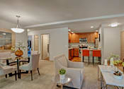 Thumbnail 5 of 32 - A bright living and dining area here at Windsong featuring beige armchairs, a glass dining table, and orange kitchen barstools. Inviting space with soft carpet, warm wood cabinetry, and an open, welcoming layout filled with light.
