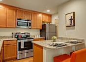 Thumbnail 6 of 32 - A cozy kitchen here at Windsong featuring warm wood cabinetry, stainless steel appliances, and bright orange barstools. Welcoming layout with a breakfast bar, clean finishes, and inviting, functional design.