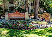 Thumbnail 32 of 32 - A community entrance sign here at Windsong featuring a warm, welcoming monument sign surrounded by colorful flowers, decorative rocks, mature trees, and lush green landscaping with apartment buildings in the background.
