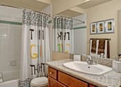 Thumbnail 11 of 32 - A clean bathroom here at Windsong featuring a granite vanity, large mirror, and bright lighting. Neutral tile shower with patterned curtain and warm wood cabinetry create a comfortable, inviting space.